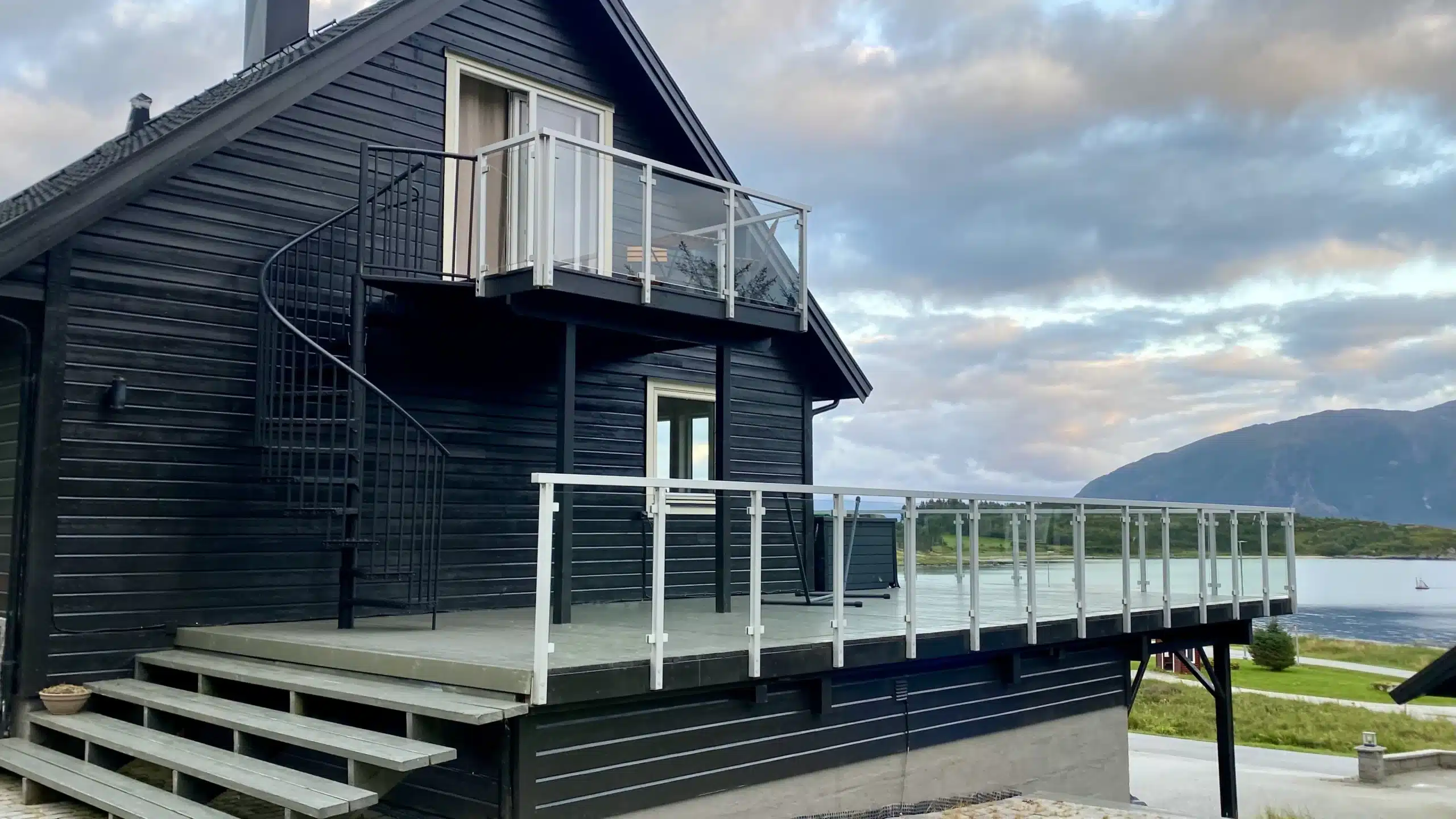 Stor träaltan med glasräcke vid svart trähus och utsikt över vatten.