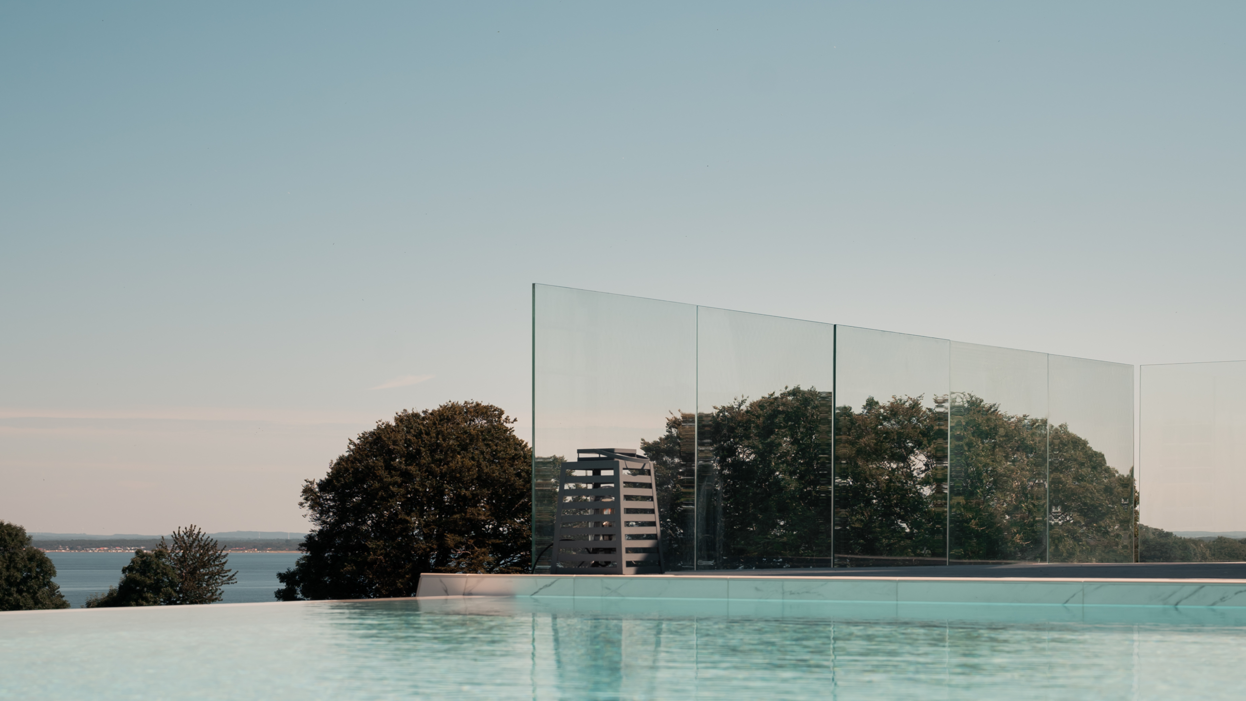 Stolpfritt glasräcke framför en pool med reflektion av träd.