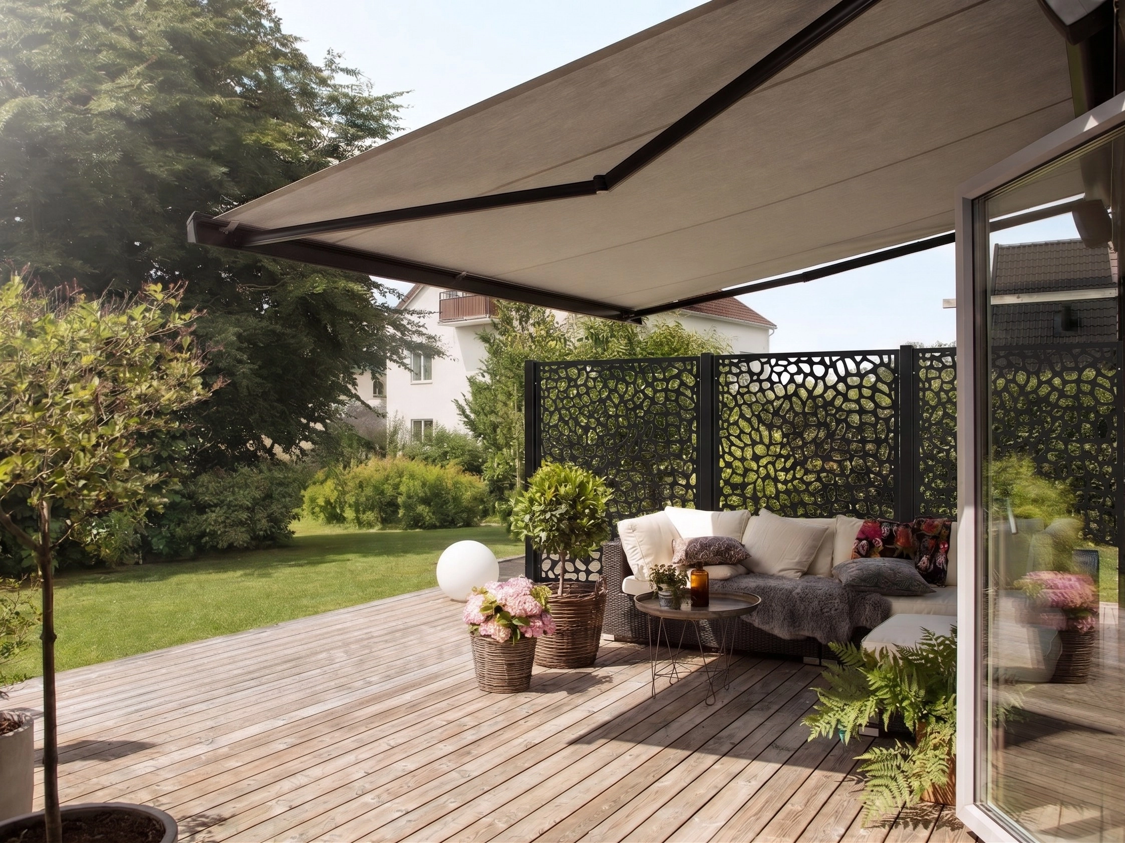 Utfälld terrassmarkis över altan med soffgrupp, krukväxter, trätrall och svart mönstrat insynsskydd i bakgrunden.