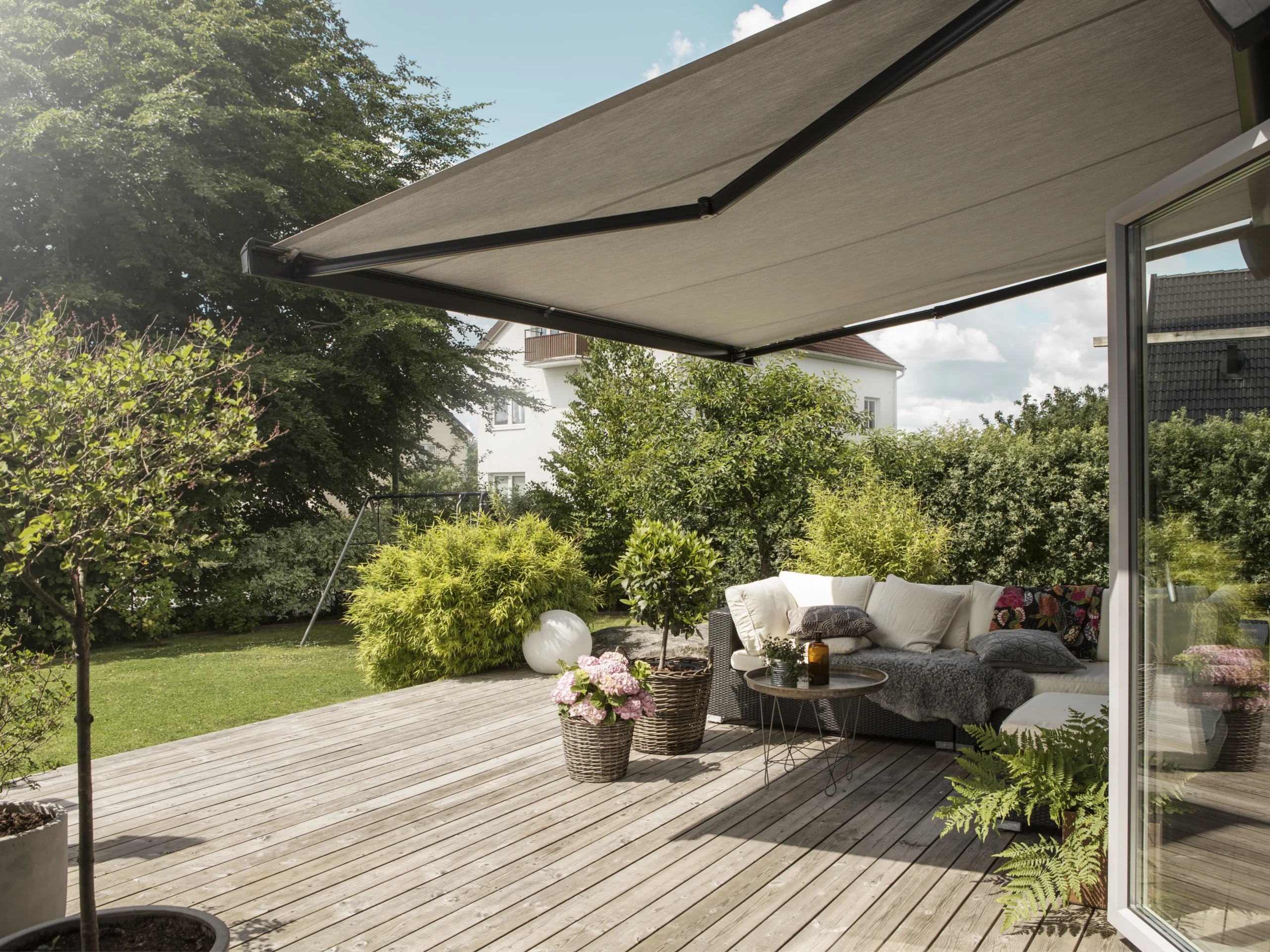 Utfälld terrassmarkis över altan med soffgrupp, krukväxter, trätrall och svart mönstrat insynsskydd i bakgrunden.