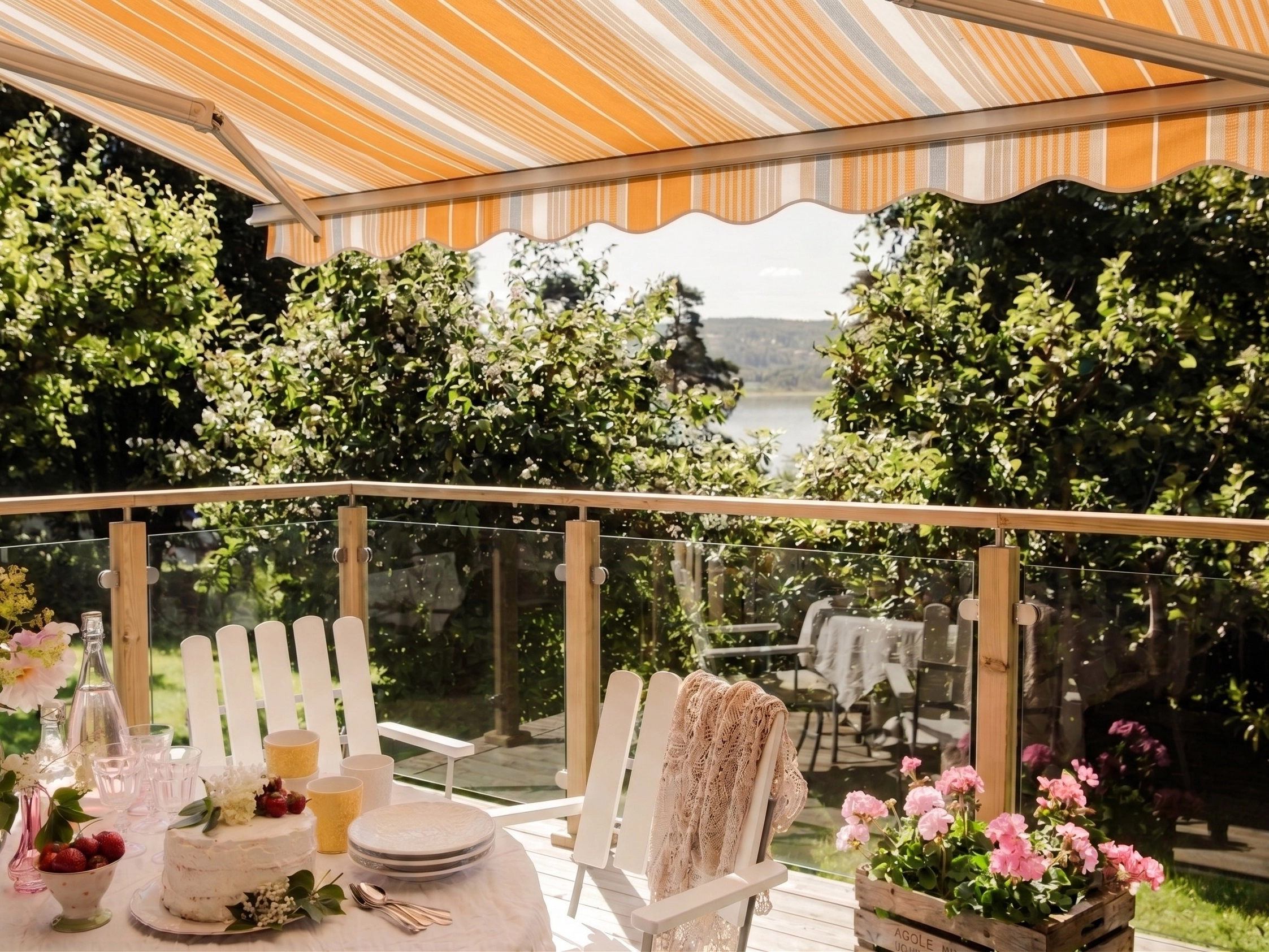 Randig terrassmarkis över balkong med glasräcke, vita stolar, dukat bord, blommor och utsikt mot vatten.