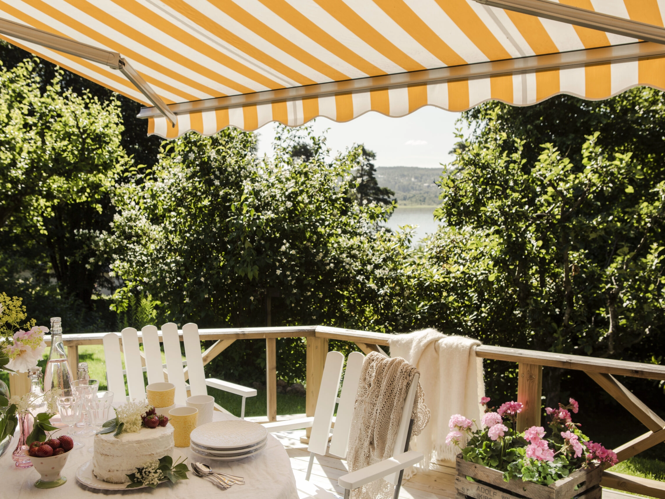 Randig terrassmarkis över balkong med glasräcke, vita stolar, dukat bord, blommor och utsikt mot vatten.