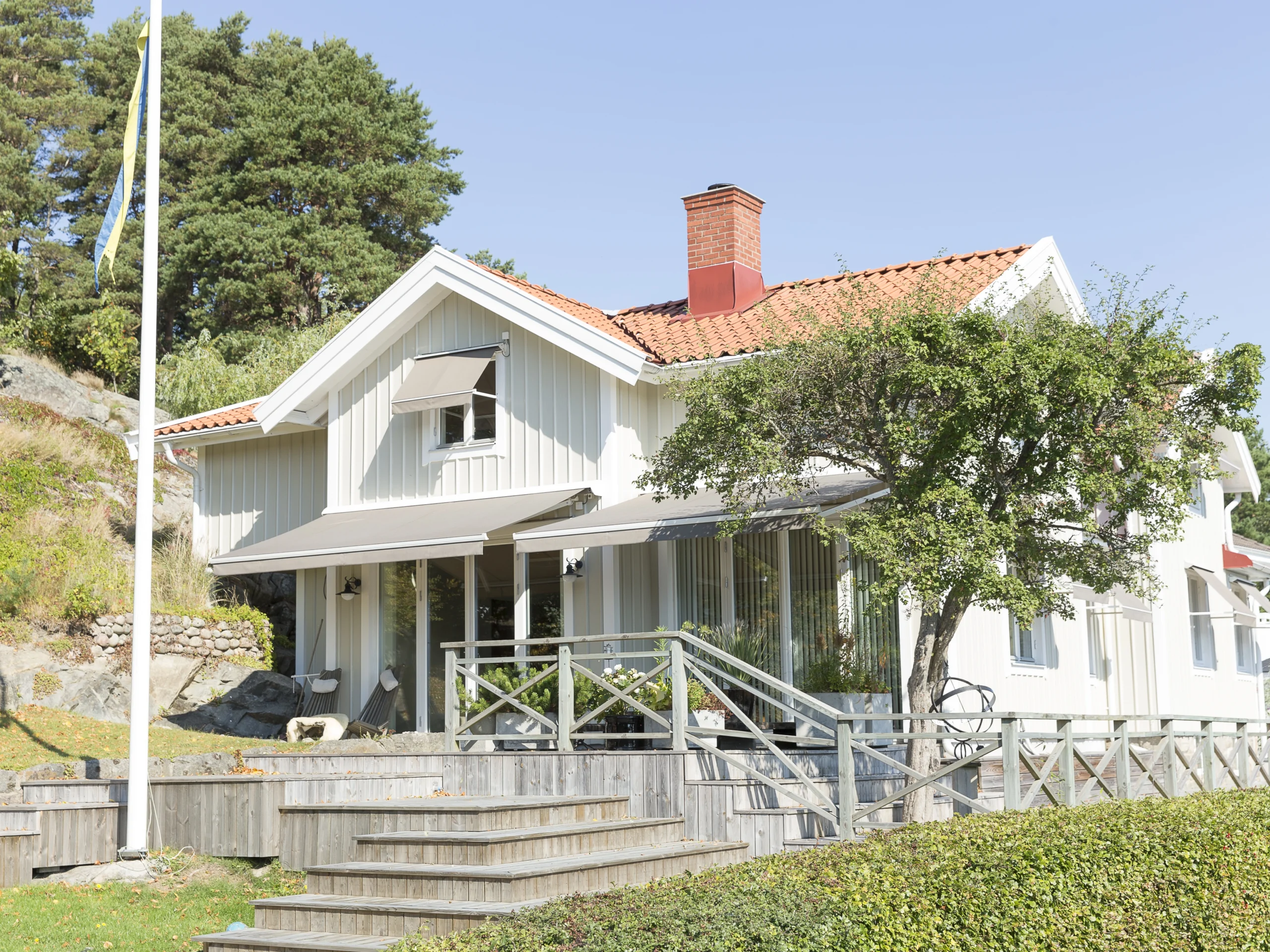 Vit villa med utfälld terrassmarkis över altan, trätrappa, glasräcke och trädgård framför huset.