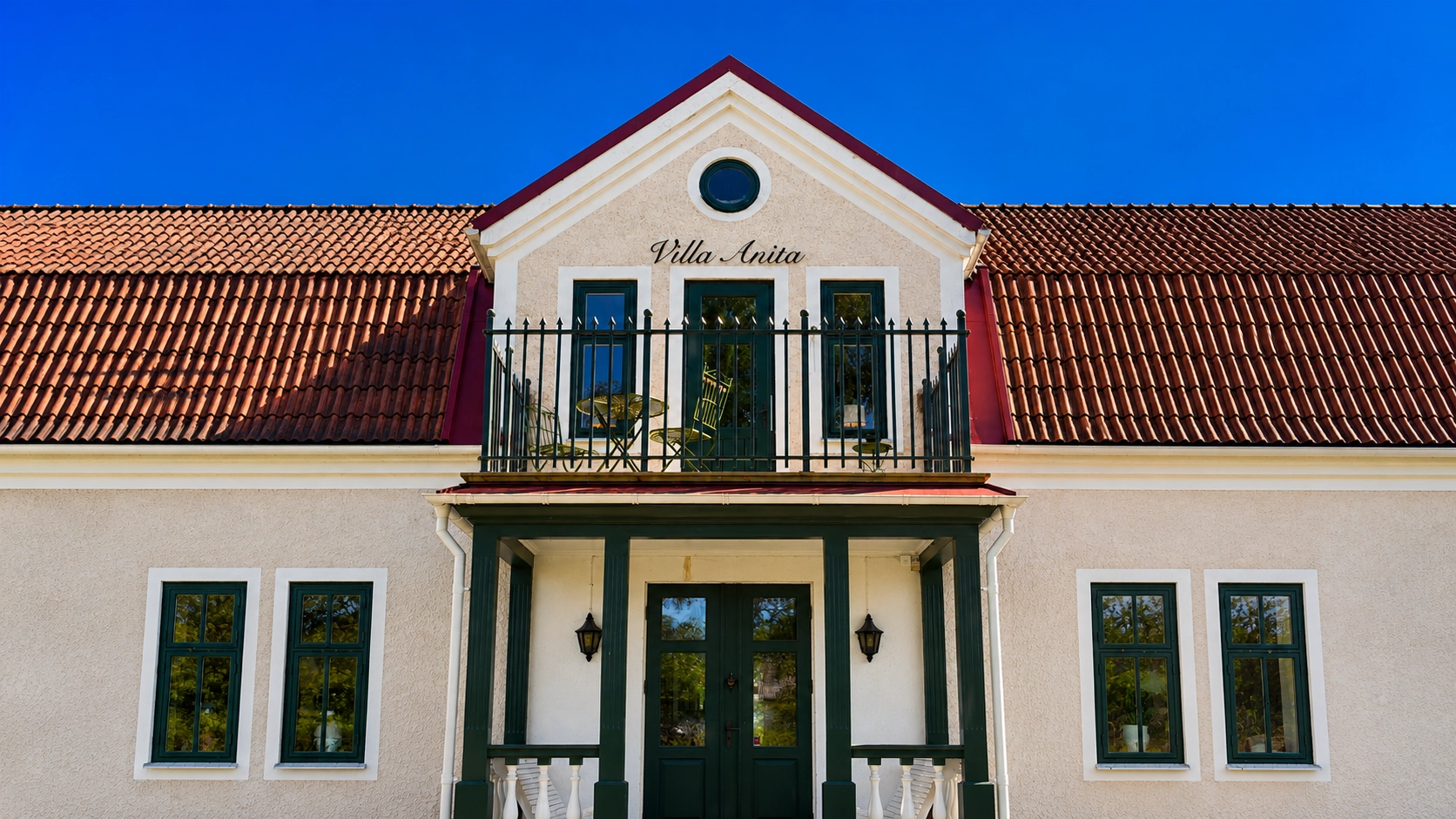 Mörkgrönt smidesräcke på balkong framför ljus villafasad med terrakottafärgat tak och gröna fönster.
